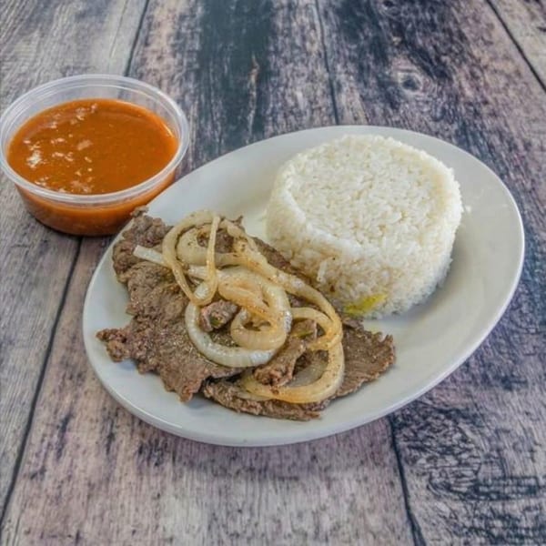 Steak with grilled onions, white rice, and a side of red sauce on a wooden table.