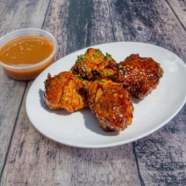 Four glazed chicken wings on a white plate with a side of dipping sauce on a wooden table.