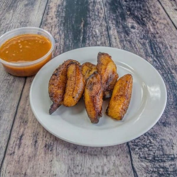 A white plate of golden brown fried plantains with a side of orange dipping sauce.